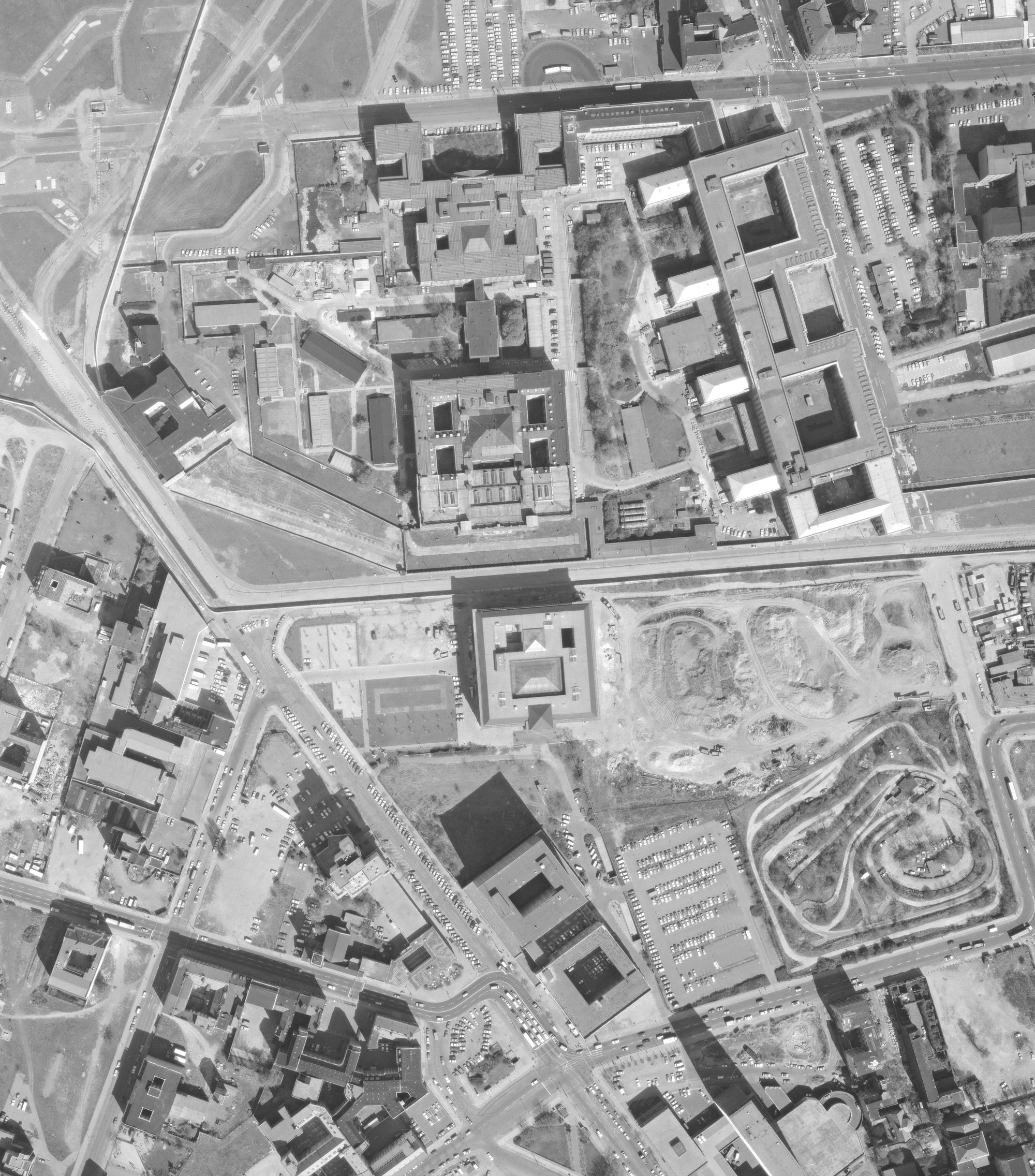 Luftbild: In der Mitte eine Brachfläche. Westlich der Martin-Gropius-Bau, südlich das Autodrom. Nördlich die Berliner Mauer, dahinter Abgeordnetenhaus und Reichsluftfahrtministerium. Grabungsort nahe dem Gropius-Bau, künftige Fundorte an der Mauer.