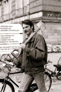 Mann mit Spaten über der Schulter, der vor einem historischen Gebäude geht. Im Hintergrund ein Schild mit der Aufschrift: „Hier befanden sich die Folterzellen der Geheimen Staatspolizei.“, darunter eine englische und französische Übersetzung.