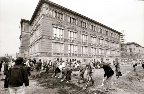 Menschen graben mit Spaten auf einem offenen Gelände vor einem großen historischen Gebäude mit stuckverzierter Fassade. Eine Seite des rechtwinkligen Grabens verläuft parallel zur Gebäudefront. Weitere Personen stehen am Rand und beobachten.