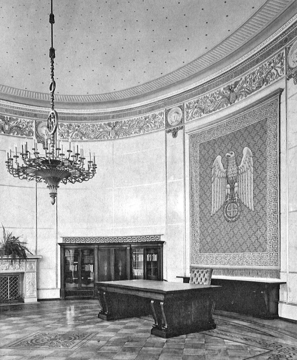 Schwarz-weiß-Fotografie auf der ein repräsentatives Amtszimmer mit großem Schreibtisch, Kronleuchter und Wandteppich mit Reichsadler abgebildet ist.
