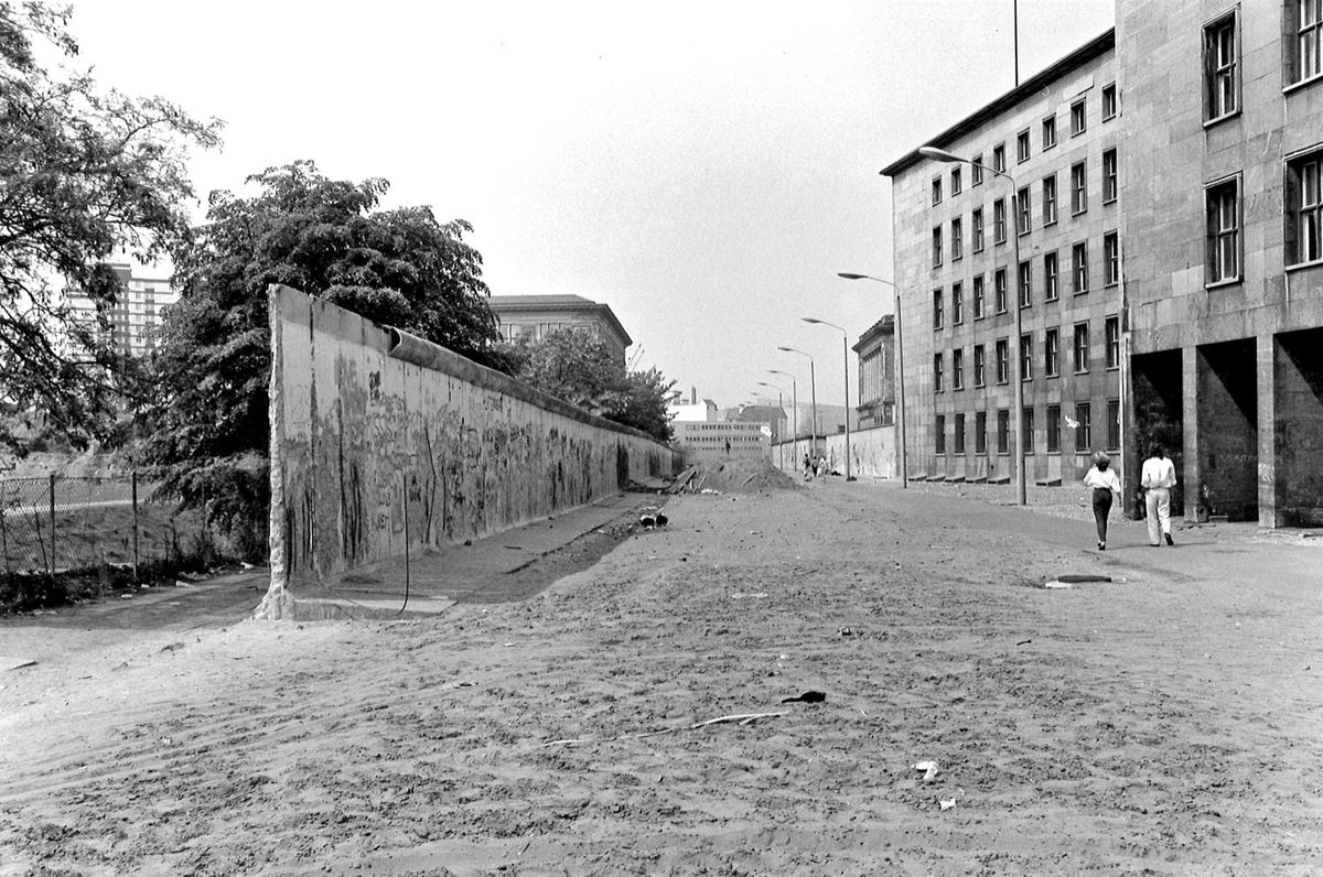 Schwarz-weiß-Fotografie: Blick in eine leere Straße entlang der Berliner Mauer. Links die Grenzmauer, rechts ein großes, geradliniges Gebäude. Zwei Personen gehen die Straße entlang, der Boden ist unbefestigt.