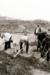 In Form eines L graben Menschen mit Spaten in der Erde eine etwa 30 cm tiefe Rinne. Im Hintergrund ein mit Gras bewachsener Erdhügel. Hinter einer Mauer ein großes Gebäude mit geradliniger Architektur.