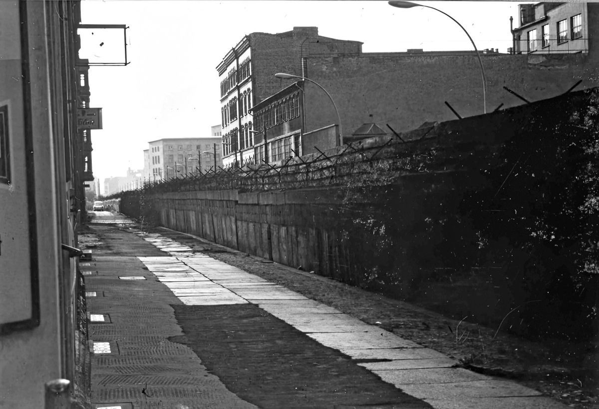 Schwarz-weiß-Fotografie einer menschenleeren Straße entlang der Berliner Mauer, flankiert von hohen Gebäuden.
