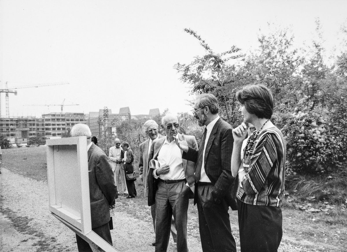 Schwarz-weiß-Fotografie: Eine Gruppe von Personen steht im Gespräch auf einer unbebauten Fläche und betrachtet eine Tafel. Im Hintergrund Baukräne und Rohbauten, rechts Büsche und Bäume.
