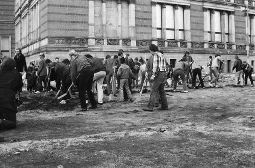 Zahlreiche Menschen graben mit Spaten auf einem unbefestigten Gelände neben einem historischen Gebäude. Sie arbeiten in L-Form, die lange Seite verläuft entlang der Gebäudefront. Links kniet eine Person mit Kamera, im Vordergrund harter Boden.