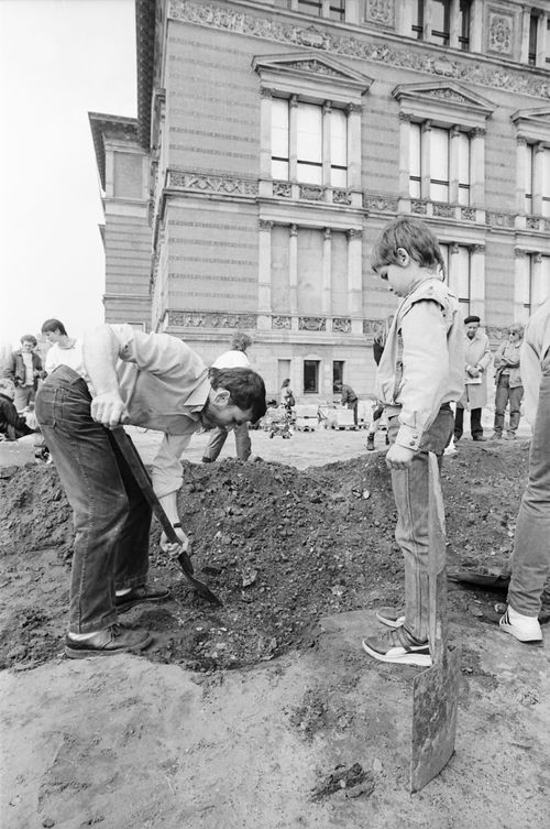 Ein Mann beugt sich mit einem Spaten über aufgewühlte Erde. Rechts daneben steht ein Kind auf einem kleinen Steinquader und schaut hinab. Im Hintergrund ein großes historisches Gebäude mit verzierter Fassade und weiteren Personen.