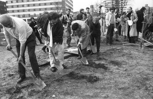 Vier Personen beginnen entlang einer in die Erde geritzten Markierung zu graben. Im Hintergrund stehen Menschen und beobachten das Geschehen. Drei von ihnen halten Zettel in der Hand und schauen aufmerksam zu.