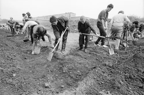 Eine Gruppe gräbt mit Spaten entlang eines rechtwinkligen, flachen Grabens. Die Grabenecke liegt unten im Vordergrund, mehrere Personen arbeiten gebeugt. Die Reihe setzt sich nach rechts und links fort.
