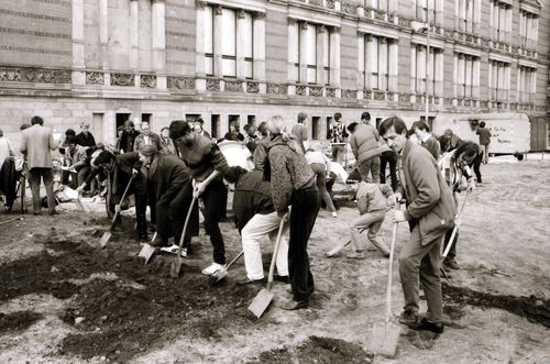 Eine Gruppe von Menschen, darunter auch ein Kind, steht vor einem historischen Gebäude und gräbt nebeneinander mit Spaten in dunkler Erde. Einige beugen sich vor, andere schauen pausierend auf. Im Hintergrund ein Bauwagen und beobachtende Personen.