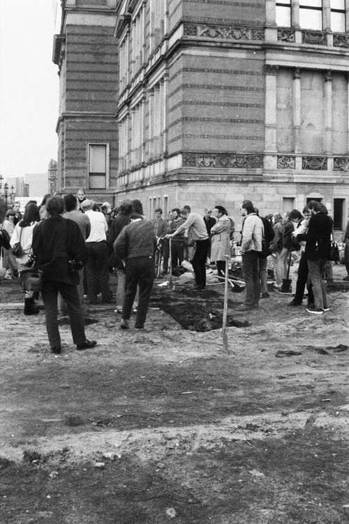 Menschen stehen dicht beieinander auf einem unbefestigten Gelände neben einem historischen Gebäude. Zwischen ihnen verläuft eine gerade Linie aufgeworfener Erde. Der Boden im Vordergrund ist hart, ein Spaten steckt in der Erde.