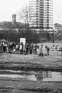 Menschen stehen und graben auf einem unbefestigten Gelände neben einem historischen Gebäude. Im Vordergrund eine große Pfütze mit Reifenspuren, im Hintergrund zwei moderne mehrstöckige Gebäude und Bäume.