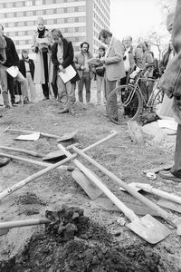 Mehrere Spaten liegen kreuz und quer auf dem Boden. Dahinter steht eine Gruppe mit ernsten Gesichtern um eine Frau, die auf einem flachen Erdhügel etwas vorliest. Daneben eine Frau mit verschränkten Armen und gesenktem Blick. Im Hintergrund ein Hochhaus.