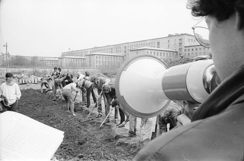 Rechts im Vordergrund eine Person mit Brille, die in ein Megafon spricht. Links im Bildrand sind Textblätter zu sehen. Eine lange Reihe Menschen gräbt konzentriert einen schmalen Graben. Im Hintergrund ein massives Gebäude mit klar gegliederter Fassade.