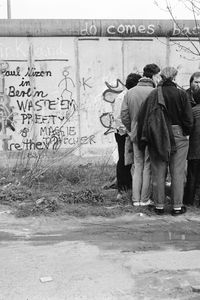 Eine kleine Gruppe von Personen steht dicht beisammen vor der Berliner Mauer, die mit zahlreichen Graffiti besprüht ist. Das Graffiti zeigt unterschiedliche Schriftzüge und ein Gesicht mit Hut und Brille. Vor der Mauer eine Wasserpfütze und niedriges Gras
