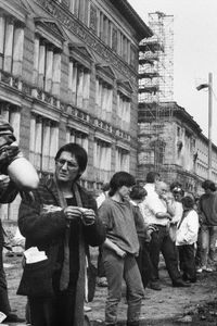 Vor einem historischen Gebäude stehen Menschen am Rand eines aufgewühlten Erdabschnitts. Im Vordergrund dreht ein Mann eine Zigarette. Rechts stehen Spaten und Schaufeln in den umgegrabenen Erdabschnitt gesteckt.