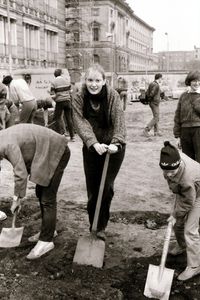 Eine Frau blickt freundlich in die Kamera und stützt einen Fuß auf einen Spaten. Neben ihr graben zwei Personen in der Erde, darunter ein Kind mit Mütze. Im Hintergrund Baugerüste und historische Gebäude.