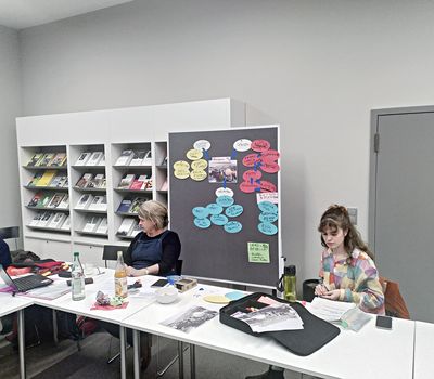 Zwei Frauen sitzen an einem Tisch mit Arbeitsmaterialien, im Hintergrund eine Pinnwand mit farbigen Zetteln, die ein thematisches Cluster um ein Foto bilden.