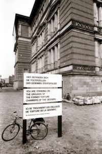Ein Schild vor einer historischen Gebäudeecke trägt den Hinweis: „Hier befanden sich die Folterzellen der Geheimen Staatspolizei.“ Darunter Übersetzungen auf Englisch und Französisch. Links lehnt ein Fahrrad am Schild.