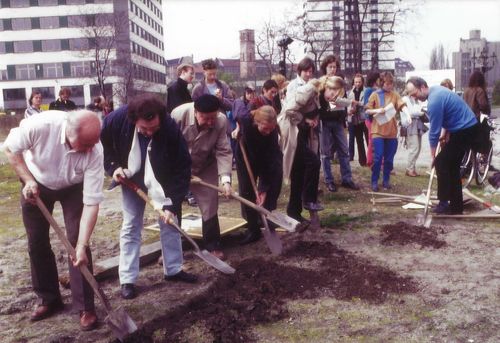 Sechs Personen graben mit Spaten entlang einer geraden Linie, eine siebte im rechten Winkel dazu. Der Graben ist flach, sie haben gerade begonnen. Menschen beobachten das Geschehen, einige halten Zettel.