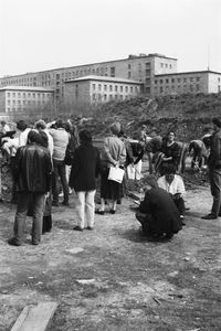Menschen graben auf einer offenen Fläche. Vor den Grabenden stehen oder hocken andere, die das Geschehen beobachten. Hinter der Gruppe ein niedriger Erdhügel, dahinter ragt ein großer, mehrflügeliger Gebäudekomplex mit schlichter Fassade hervor.