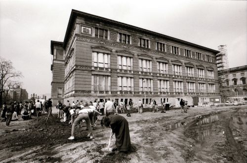 Vor einem historischen Gebäude mit stuckverzierter Fassade graben Menschen mit Spaten eine rechtwinklige Rinne. Rechts im Bild große Pfützen und deutliche Reifenspuren im unebenen Boden.