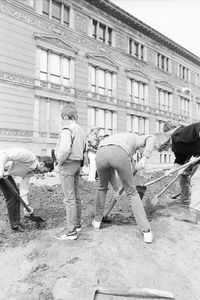 Mehrere Personen graben mit Spaten an einem flachen Graben vor einem historischen Gebäude mit verzierter Fassade. Im Zentrum steht ein Kind aufrecht zwischen den Grabenden und blickt auf die aufgewühlte Erde.