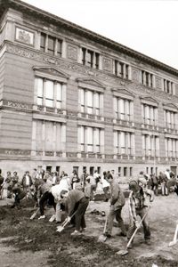 Menschen graben mit Spaten auf einem offenen Gelände vor einem großen historischen Gebäude mit stuckverzierter Fassade. Eine Seite des rechtwinkligen Grabens verläuft parallel zur Gebäudefront. Weitere Personen stehen am Rand und beobachten.