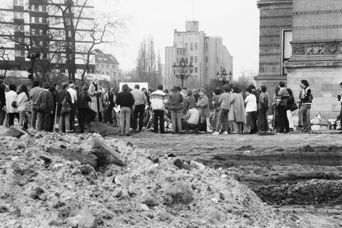 Eine große Menschengruppe steht auf einem unbefestigten Gelände neben einem historischen Gebäude. Im Vordergrund ein Hügel aus aufgeworfener Erde und Reifenspuren, im Hintergrund Bäume und mehrstöckige Gebäude.
