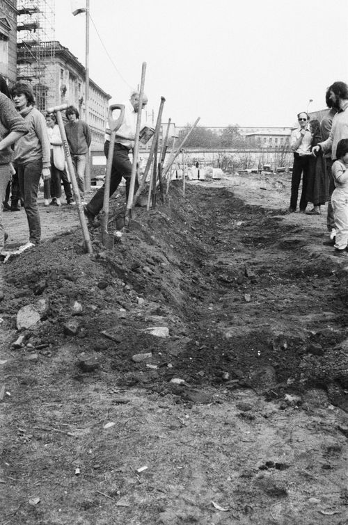 Ein flacher Graben mit aufgeworfener Erde verläuft mittig durchs Bild. In der angehäuften Erde stecken Spaten und Schaufeln. Links und rechts des Grabens stehen Menschen. Im Hintergrund ein historisches Gebäude und die Berliner Mauer.