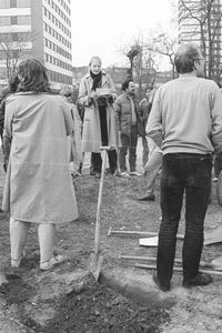 Auf einem offenen Gelände stehen zahlreiche Menschen in einem Kreis. In der Mitte liest eine Frau von einem Zettel. Spaten liegen auf dem Boden. Im Vordergrund ist ein Kind zu sehen, im Hintergrund ein Fahrrad und eine mehrsprachige Hinweistafel.
