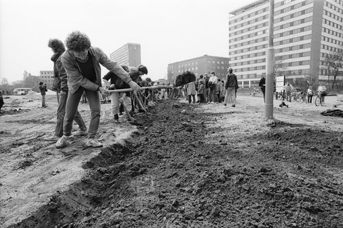 Mehrere Menschen graben in einer langen Reihe mit Spaten einen flachen Graben in aufgewühlter Erde. Rechts moderne mehrstöckige Gebäude. Im Hintergrund einige zuschauende Personen.