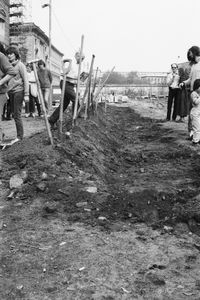 Ein flacher Graben mit aufgeworfener Erde verläuft mittig durchs Bild. In der angehäuften Erde stecken Spaten und Schaufeln. Links und rechts des Grabens stehen Menschen. Im Hintergrund ein historisches Gebäude und die Berliner Mauer.