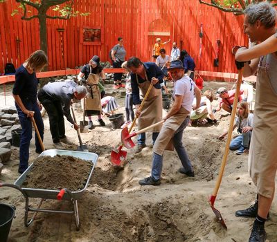 In einem umzäunten Außenbereich mit roter Holzwand graben mehrere Menschen mit Schaufeln und einer Schubkarre in sandigem Boden. Einige tragen Schürzen und Arbeitshandschuhe. Kinder und Erwachsene beobachten oder helfen mit.